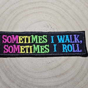 May include: Black embroidered patch with the text "SOMETIMES I WALK, SOMETIMES I ROLL" in pink, yellow, green, and blue thread.
