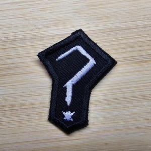 Puede incluir: Parche de tela negro con un símbolo bordado en blanco que se asemeja a un número siete estilizado.