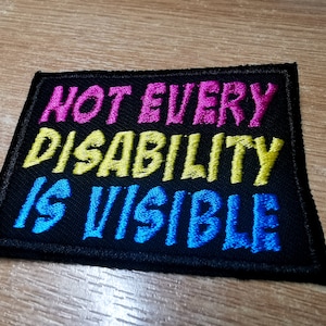 Op de afbeelding: Zwarte geborduurde patch met de tekst "Not Every Disability Is Visible" in roze, gele en blauwe letters.