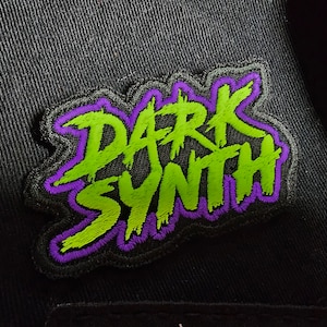 Puede incluir: Un parche bordado verde y morado con el texto "DARK SYNTH" en una fuente de graffiti estilizada.