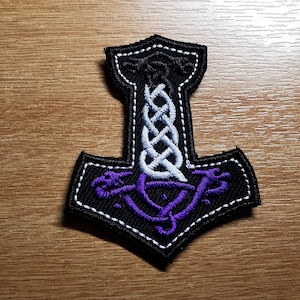 May include: Black embroidered patch with a white and purple design of a Norse hammer with a knotwork pattern.