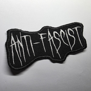 May include: Black embroidered patch with the words "ANTI-FASCIST" in white, stylized lettering. The patch has a unique, irregular shape and a black border. The text is in a bold, slightly distressed font.