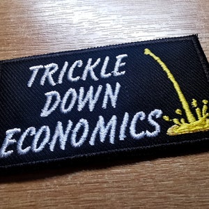 May include: Black embroidered patch with white text that reads "Trickle Down Economics". A yellow graphic of a dripping liquid is on the right side of the patch.