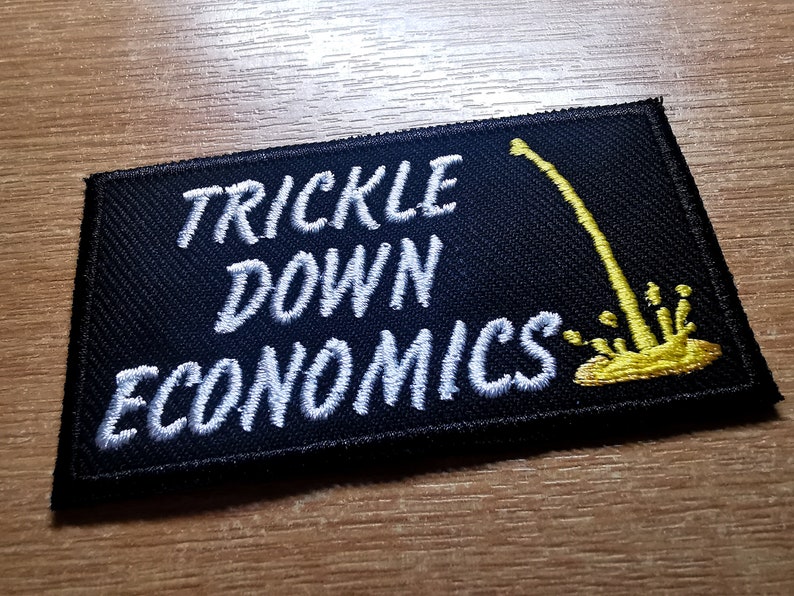 May include: Black embroidered patch with white text that reads "Trickle Down Economics". A yellow graphic of a dripping liquid is on the right side of the patch.