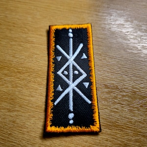 May include: A black embroidered patch with a white geometric design. The patch has an orange border and is sewn onto a wooden surface.