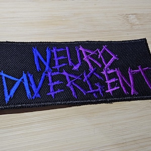 Pode incluir: Um remendo de tecido preto com as palavras "Neuro Divergent" bordadas em fio azul e roxo.