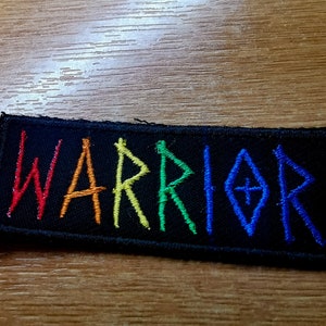 Könnte beinhalten: Ein schwarzer Stoff-Patch mit dem Wort "WARRIOR" in Regenbogenfarben gestickt. Die Buchstaben sind stilisiert und haben eine leicht gezackte Form.