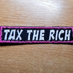 Puede incluir: Un parche de tela negro con costuras rosas alrededor de los bordes. El parche dice "TAX THE RICH" en letras blancas.
