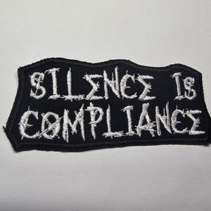 Puede incluir: Parche bordado negro con la frase "SILENCE IS COMPLIANCE" en blanco. El texto está escrito en un estilo especial. El parche tiene un borde irregular y está diseñado para coserlo en la ropa.
