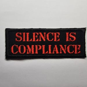 Może przedstawiać: Czarna prostokątna naszywka z wyhaftowanym czerwonym napisem "SILENCE IS COMPLIANCE". Naszywka ma czarną obwódkę i jest umieszczona na białym tle. Tekst jest pogrubiony, czcionką bezszeryfową.