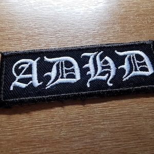 Op de afbeelding: Zwarte geborduurde patch met witte tekst "AD/HD".
