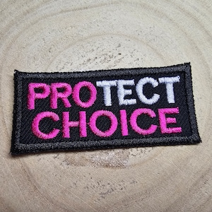 Puede incluir: Parche bordado negro con las palabras "PROTECT CHOICE" en letras blancas y rosas.