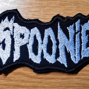 Könnte beinhalten: Ein blauer gestickter Aufnäher mit dem Wort "SPOONIE" in einer stilisierten Schriftart auf schwarzem Hintergrund.
