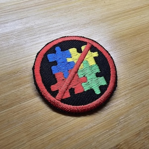 Könnte beinhalten: Gestickter Aufnäher mit einem schwarzen Kreis und rotem Rand. Im Inneren werden bunte Puzzleteile von einer roten Linie durchgestrichen. Die Puzzleteile sind blau, gelb und grün. Der Aufnäher liegt auf einer Holzoberfläche.