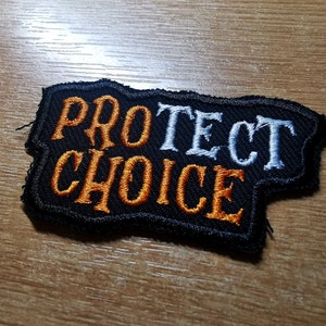 Könnte beinhalten: Ein schwarzer gestickter Aufnäher mit den Worten "PROTECT CHOICE" in weißer und orangefarbener Schrift.