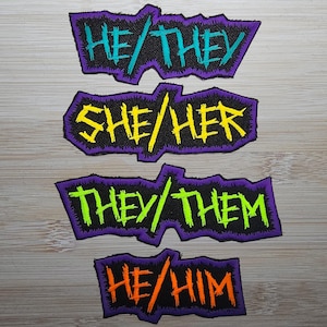 May include: Four iron-on patches with different pronoun combinations. The patches are black with neon green, yellow, and blue lettering. The text on the patches reads "He/They", "She/Her", "They/Them", and "He/Him".