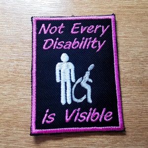 Può includere: Toppa ricamata nera con cuciture rosa. La toppa presenta una silhouette bianca di due persone, una in sedia a rotelle, e il testo "Not Every Disability is Visible".