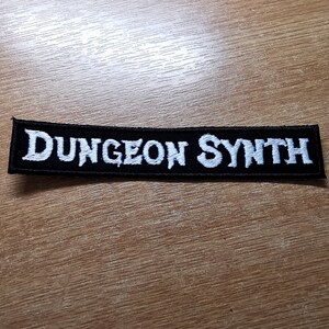 May include: Black embroidered patch with white text that reads "Dungeon Synth".