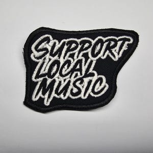 Può includere: Toppa nera con testo bianco ricamato che recita "SUPPORT LOCAL MUSIC". La toppa ha un bordo nero e uno sfondo bianco. Probabilmente è per abbigliamento o accessori.