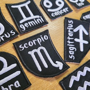 Op de afbeelding: Zwarte stoffen patches geborduurd met witte dierenriem symbolen en namen. De patches hebben verschillende vormen, waaronder een schild en een gebogen banner. Dierenriemtekens zijn onder meer Tweelingen, Schorpioen en Boogschutter.