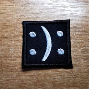 Può includere: Una toppa quadrata nera con un'emoticon sorridente capovolta ricamata in bianco.