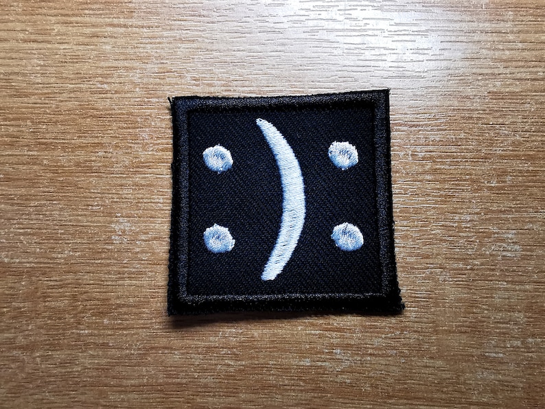 May include: A black square patch with a white embroidered upside-down smiley face.
