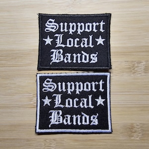 May include: Two black iron-on patches with white embroidery. The patches read "Support Local Bands" with a star on either side of the word "Local".