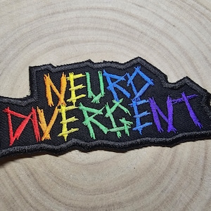 Puede incluir: Un parche de tela negro con las palabras "Neuro Divergent" bordadas en colores arcoíris. Las letras están estilizadas en una fuente irregular, de estilo punk rock.
