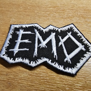 Könnte beinhalten: Weißer gestickter Aufnäher mit dem Wort "EMO" in einer gezackten, gotischen Schrift auf schwarzem Hintergrund.