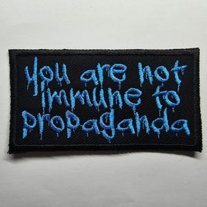 May include: Black rectangular patch with blue embroidered text that reads "You are not immune to propaganda." The text has a dripping effect. The patch has a black border and is likely made of fabric.