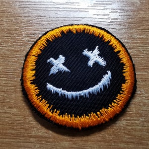 May include: A black embroidered smiley face patch with white eyes and a white smile. The patch has an orange border.