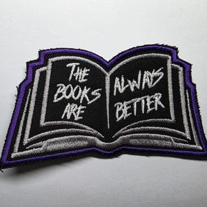 May include: Embroidered patch in the shape of an open book with the text "The Books Are Always Better". The patch has a black background with white and purple accents, and a purple border.