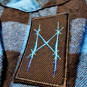 May include: A blue and black plaid flannel shirt with a black patch featuring a stylized blue 'X' design.
