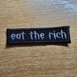 Può includere: Toppa ricamata nera con testo bianco che recita "eat the rich".