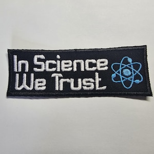 May include: A black rectangular patch embroidered with the words "In Science We Trust" in white, and a blue atom symbol. The patch has a black border and is designed for clothing or bags.