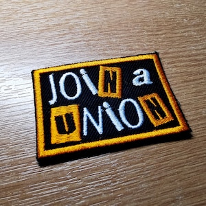 Könnte beinhalten: Ein schwarzer gestickter Aufnäher mit gelbem Rand. Der Aufnäher sagt "Join a Union" in weißen Buchstaben, wobei einige Buchstaben in gelben Kästchen sind.