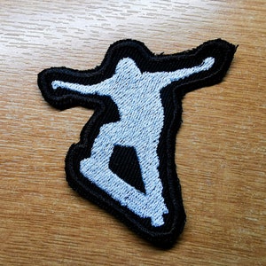 Puede incluir: Un parche bordado blanco de una persona haciendo skateboard, delineado en negro, sobre un fondo de madera.