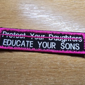 Peut inclure: Écusson brodé noir et rose avec le texte "Protect Your Daughters Educate Your Sons" en blanc.