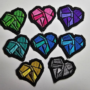 May include: Eight embroidered iron-on patches in the shape of a heart. Each heart is a different color and has a geometric pattern. The colors are green, pink, purple, teal, blue, gold, gray, and black.