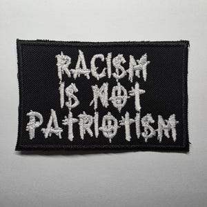 Puede incluir: Parche rectangular negro con texto bordado en blanco: «RACISM IS NOT PATRIOTISM». El texto está en una fuente estilizada. El parche tiene un borde negro y probablemente está hecho de tela.