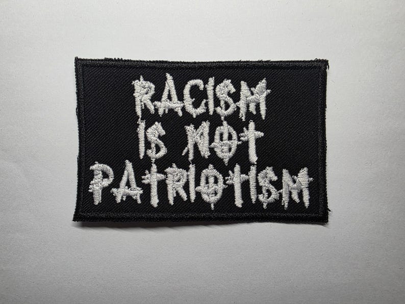 May include: Black rectangular patch with white embroidered text that reads "RACISM IS NOT PATRIOTISM". The patch has a black border and is likely made of fabric. The text is in a stylized font.