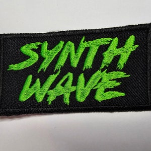 Puede incluir: Un parche de tela negro con texto bordado en verde que dice "Synth Wave".