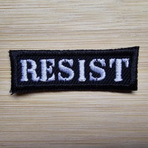 Könnte beinhalten: Schwarzer Stoff-Patch mit weiß gesticktem Text, der "RESIST" lautet.