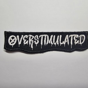 May include: Black embroidered patch with the word "OVERSTIMULATED" in white, stylized dripping font. The patch has a black border and a white background. The patch is rectangular with a wavy bottom edge.