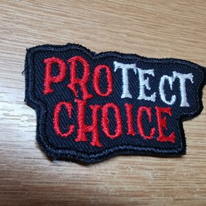 Op de afbeelding: Zwarte geborduurde patch met de woorden "PROTECT CHOICE" in witte en rode letters.