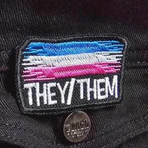 May include: A black fabric patch with a blue, white, and pink striped flag design. The text "THEY/THEM" is embroidered in white on the patch.