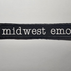 May include: A black embroidered patch with the words "midwest emo" in white, block letters. The patch is rectangular and has a stitched border. The text is centered on the patch.