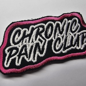 Könnte beinhalten: Gestickter Aufnäher mit dem Schriftzug "CHRONIC PAIN CLUB" in Weiß auf schwarzem Grund. Der Aufnäher hat einen pinkfarbenen Rand und ist zum Aufbügeln oder Aufnähen auf Kleidung oder Accessoires gedacht.