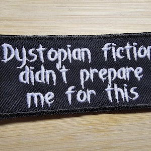 May include: Black fabric patch with white embroidered text that reads "Dystopian fiction didn't prepare me for this".