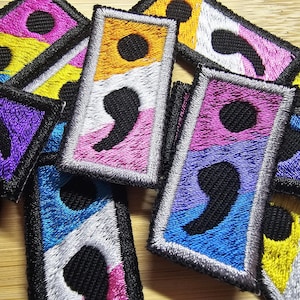 May include: A close-up of a pile of embroidered patches featuring the semicolon symbol in various colors, including yellow, purple, blue, pink, and white. The patches are made of fabric and have a black embroidered semicolon on each one.